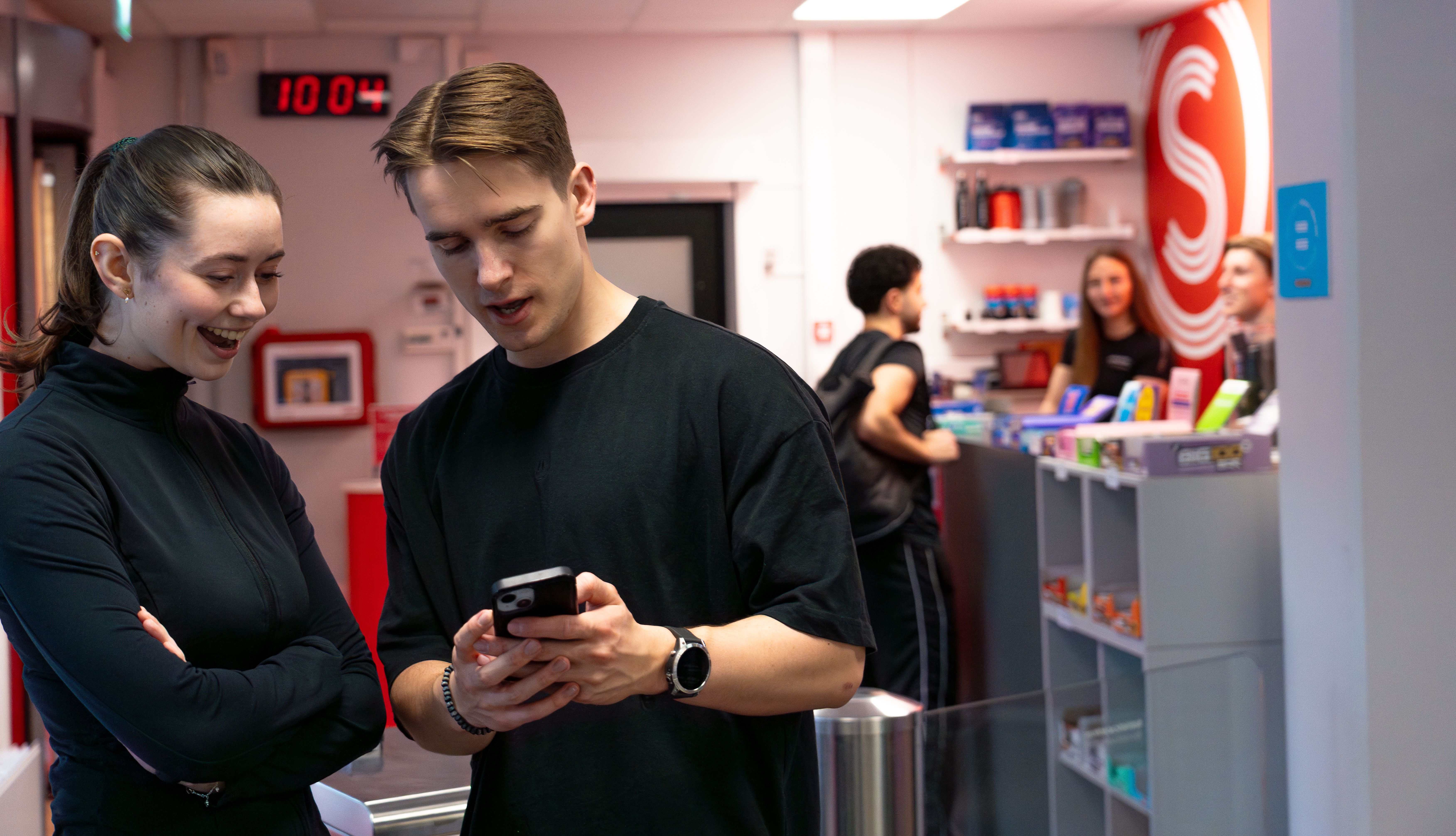 To kunder snakker sammen og ser på en telefon i resepsjonen på Athletica Centrum. 