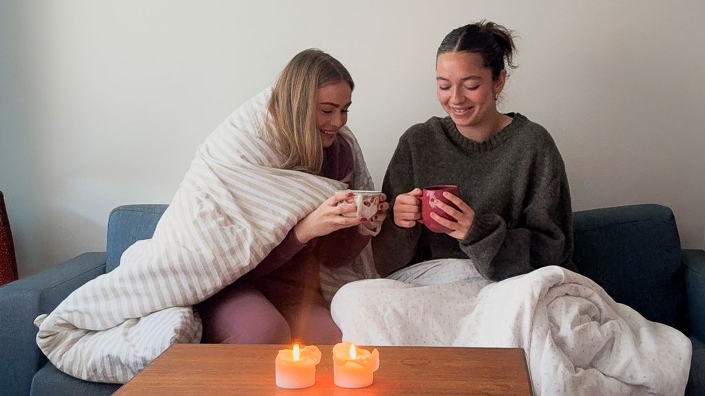 Photo of two students sitting under blankets, holding mugs of tea with lit candles in front of them.