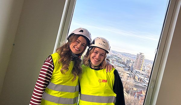 Image of two female students wearing hard hats and reflective vests, standing inside a construction site with a view of Oslo through a window behind them.