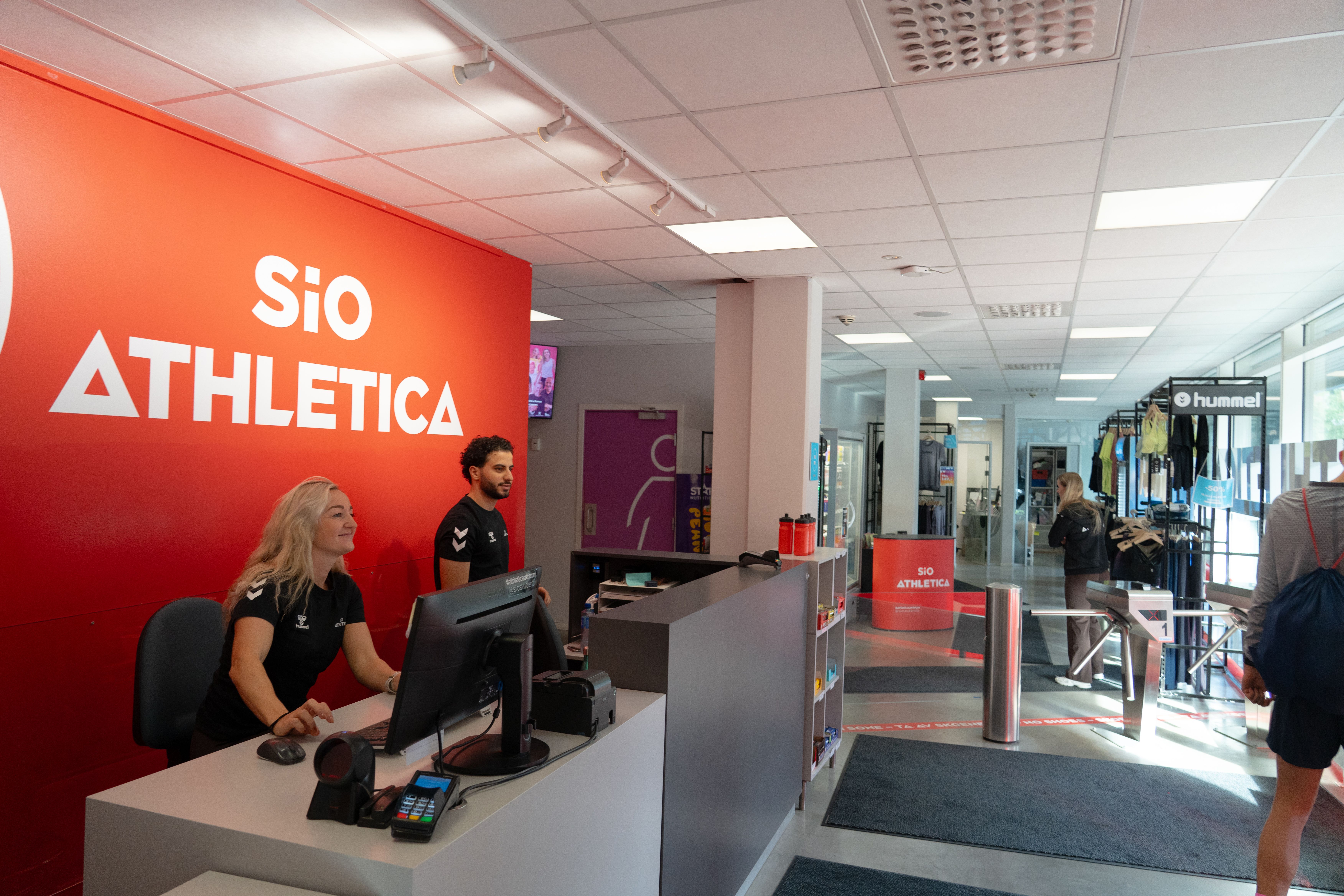 To ansatte i resepsjon- og mingleområde på Athletica Centrum med shop i bakgrunnen.