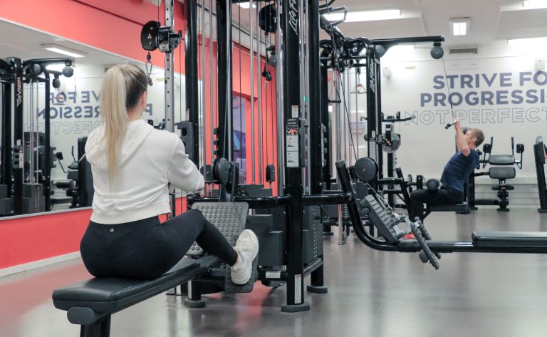 To personer som trener i treningsstudio på Athletica Blindern, en som gjør sittende roing og en som tar nedtrekk.