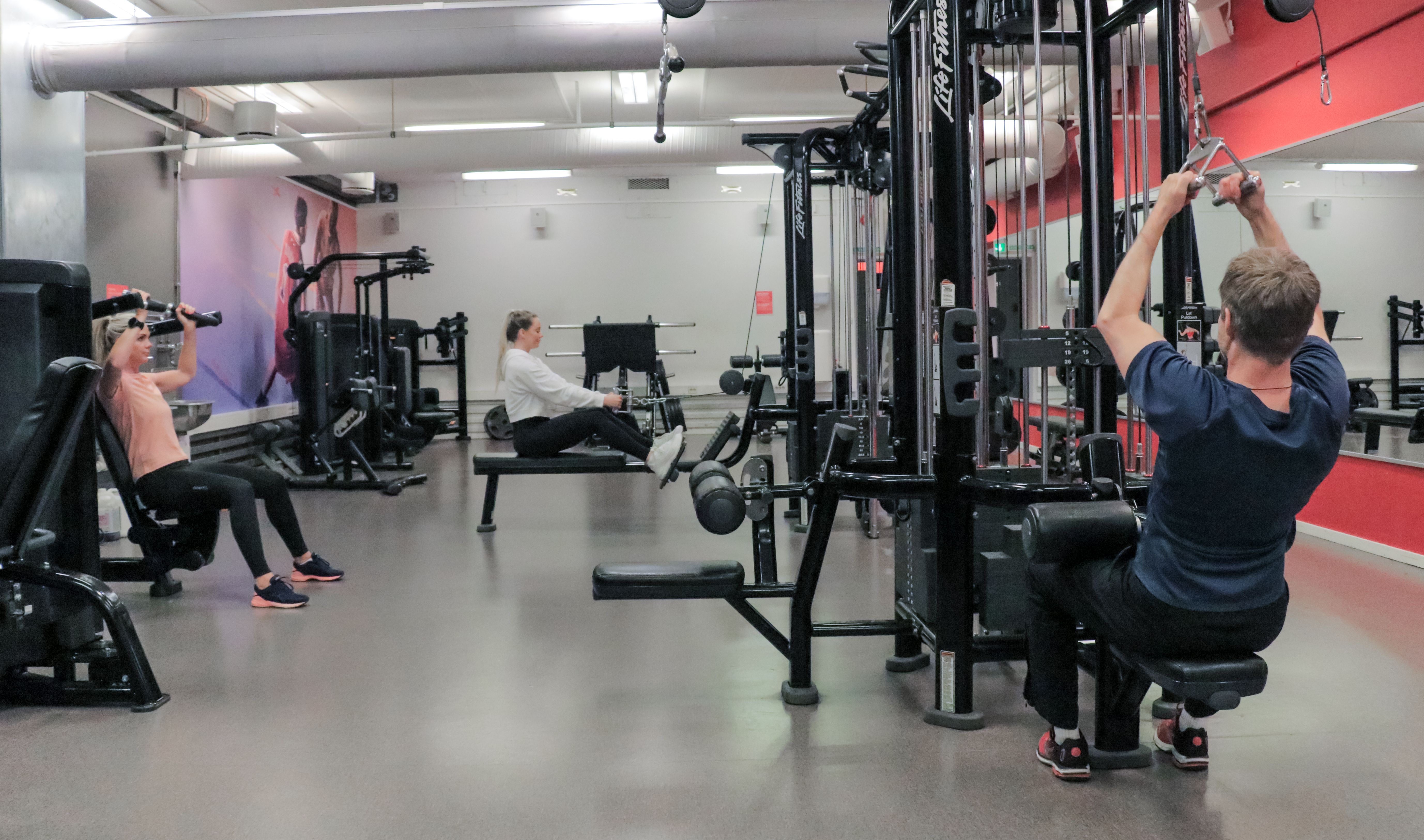 Studio på Athletica Blindern med ulike apparater og kabeltrekk, og tre personer som trener. 