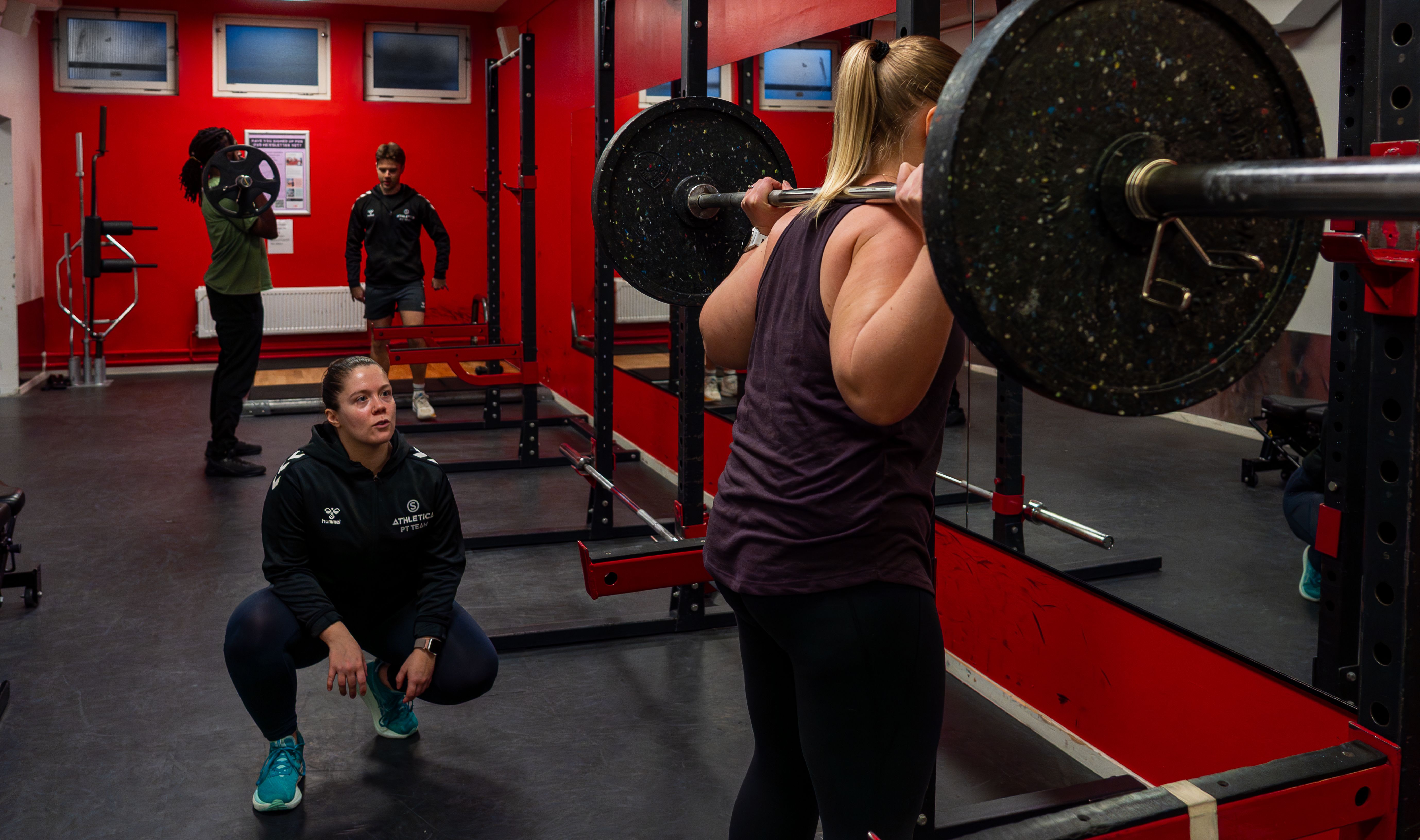 En PT som instruerer en kvinne i knebøy ved et rack, på Athletica Blindern. To andre trener i bakgrunnen.