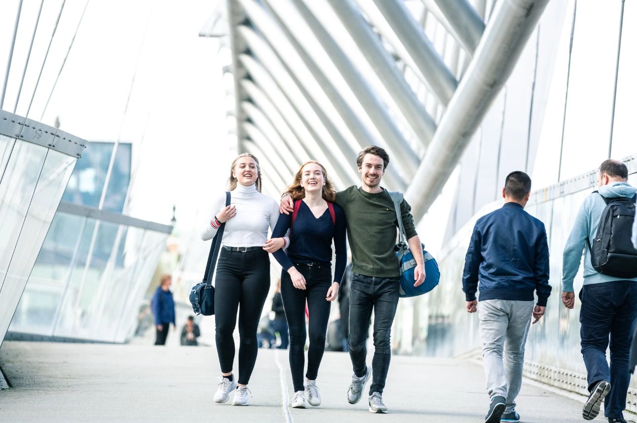 Tre nye studenter med bagger går spente sammen over en bro.