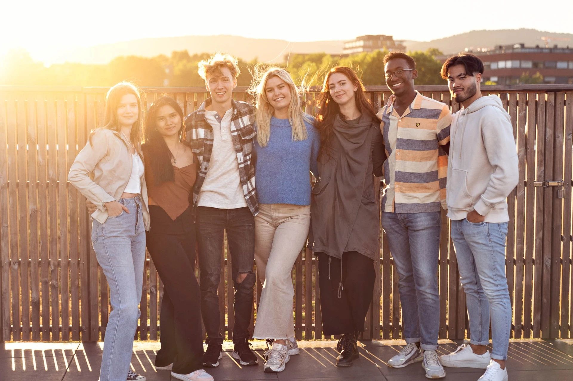 Syv studenter som står foran et gjerde med solnedgang bak seg.