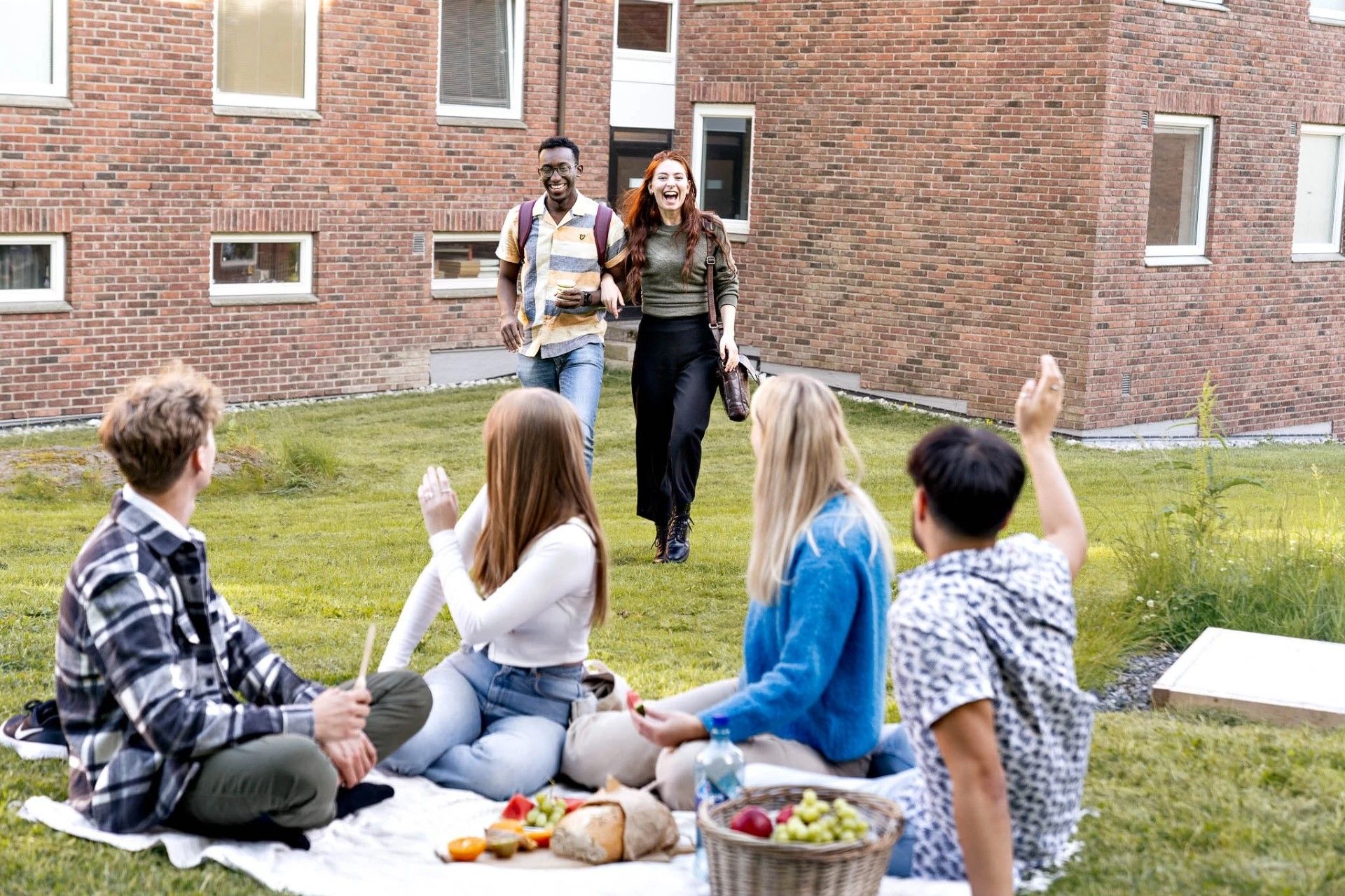 Studenter som spiser piknik foran studentbolig i murstein vinker til to glade studenter som kommer mot dem.