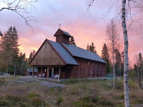Foto av nordmarkskapellet i Nordmarka i solnedgang. En hytte i tre med et kors på taket.