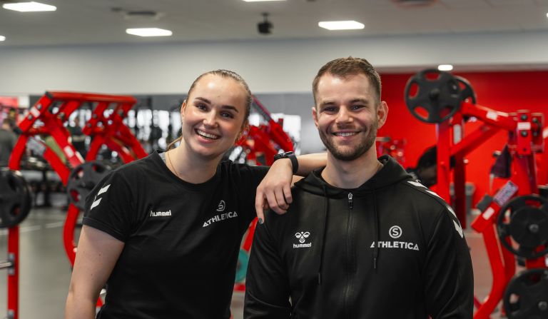 Foto av en mann og en dame som smiler foran treningsapparater som er røde og svarte.