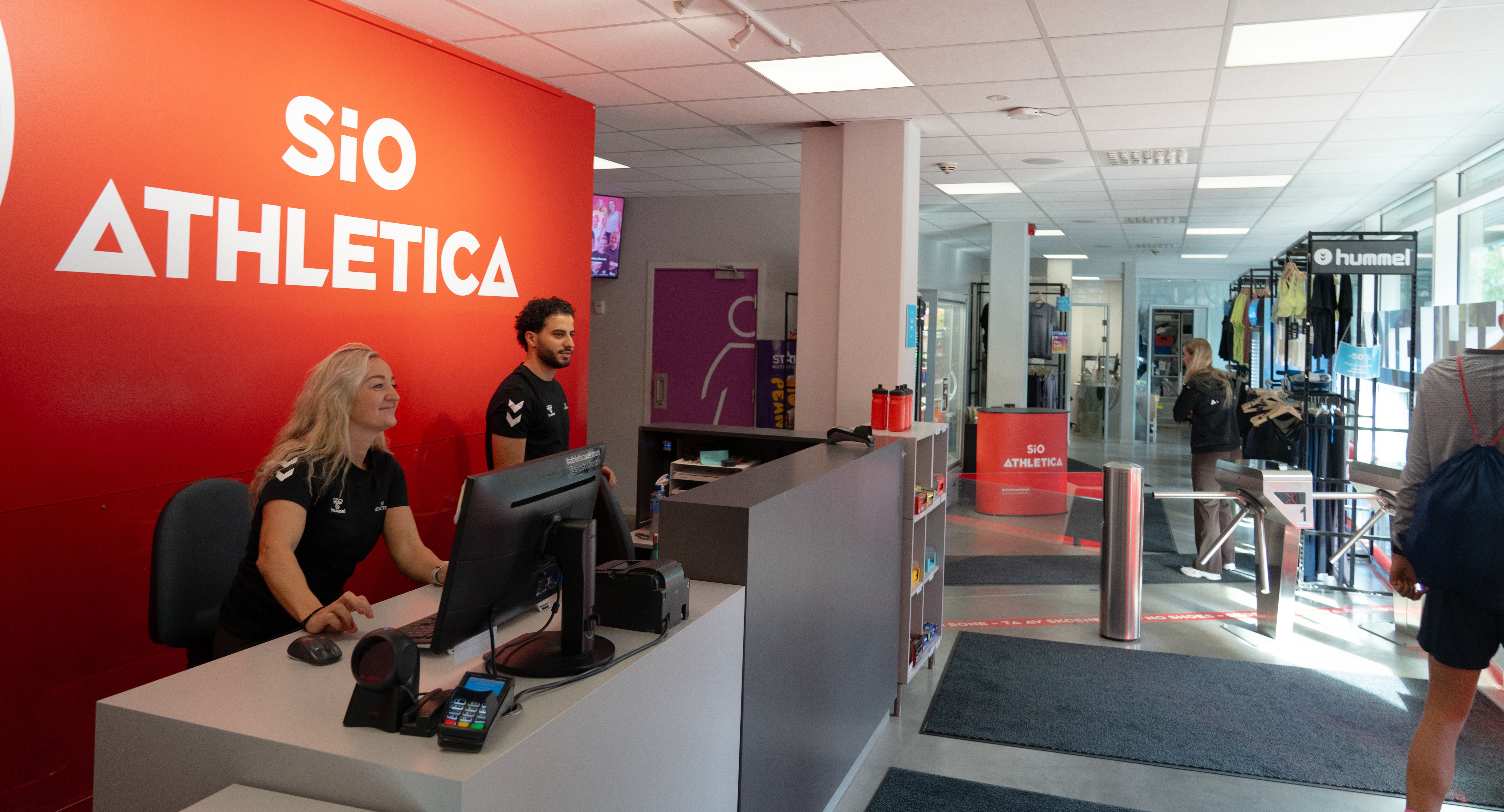 To ansatte i resepsjon- og mingleområde på Athletica Centrum med shop i bakgrunnen.