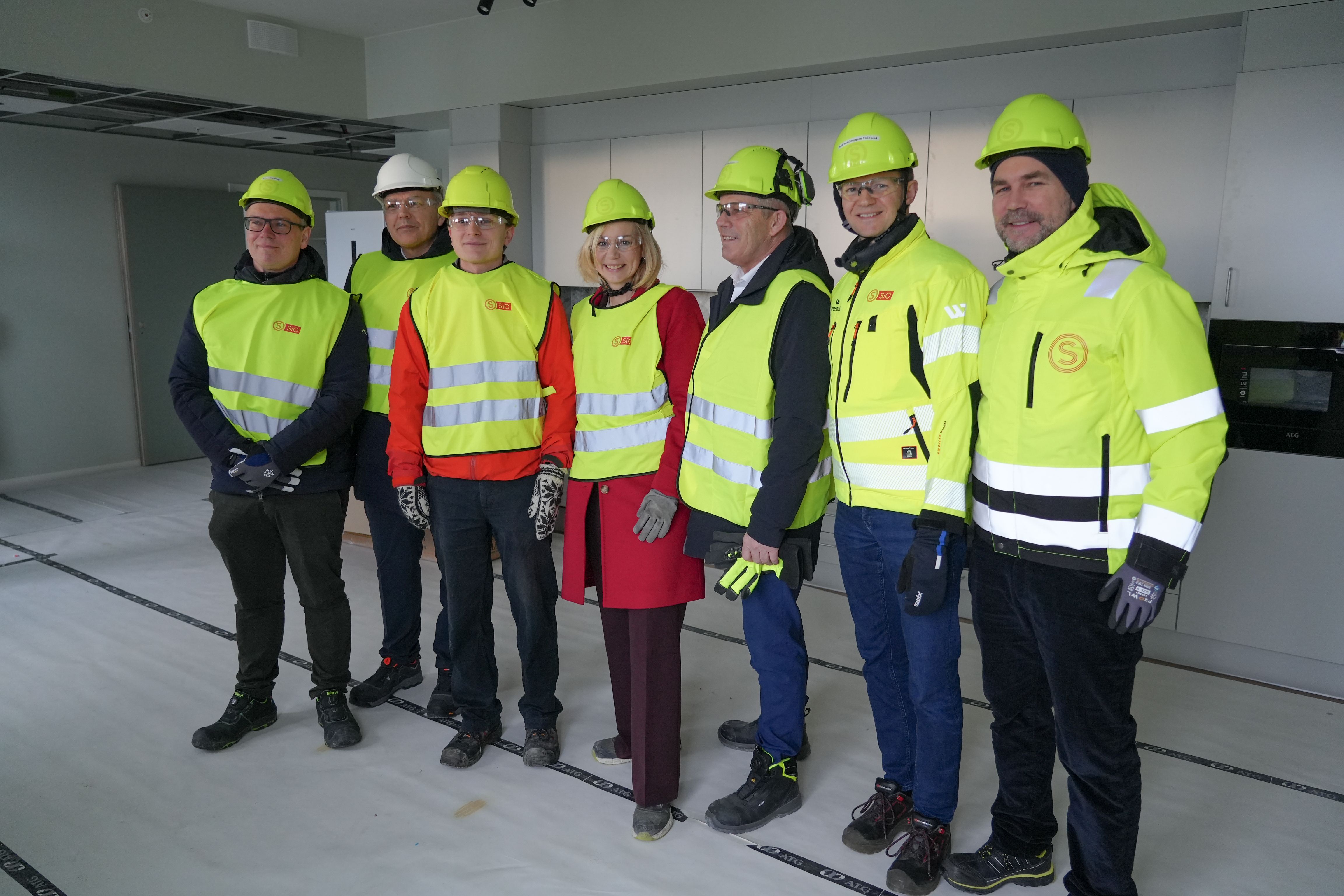 Forsknings- og høyere utdanningsminister Sigrun Aasland, kommunal- og distriktsminister Bjørnar Skjæran og administrerende direktør i Husbanken Jan Hjelle på omvisning på byggeplassen på Nordberg. Ansatte fra SiO og besøkende har på gule vester og verneutstyr og smiler til kameraet.