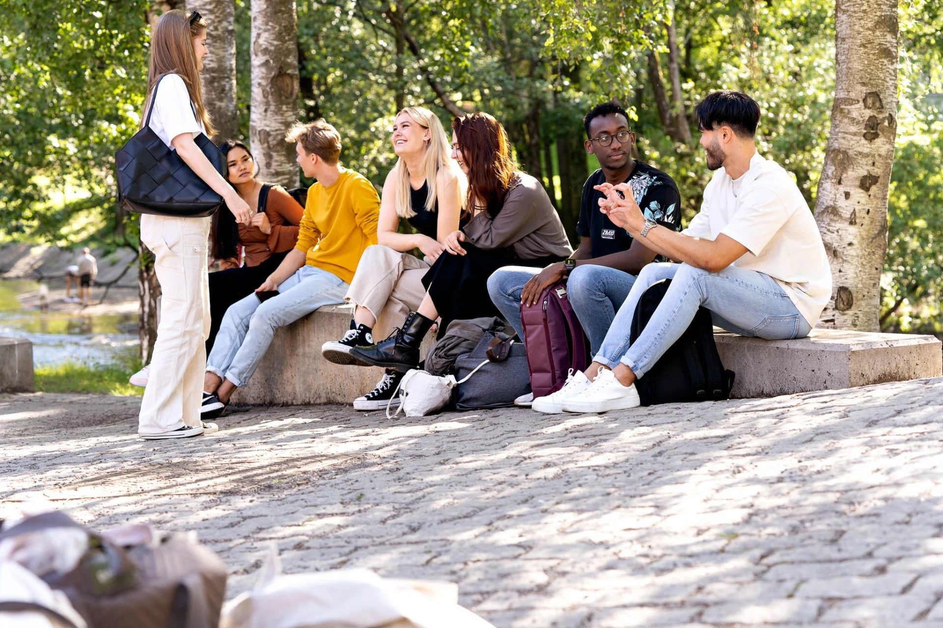 Syv studenter som sitter på en steinbenk og prater med grønne trær og Akerselva bak seg.