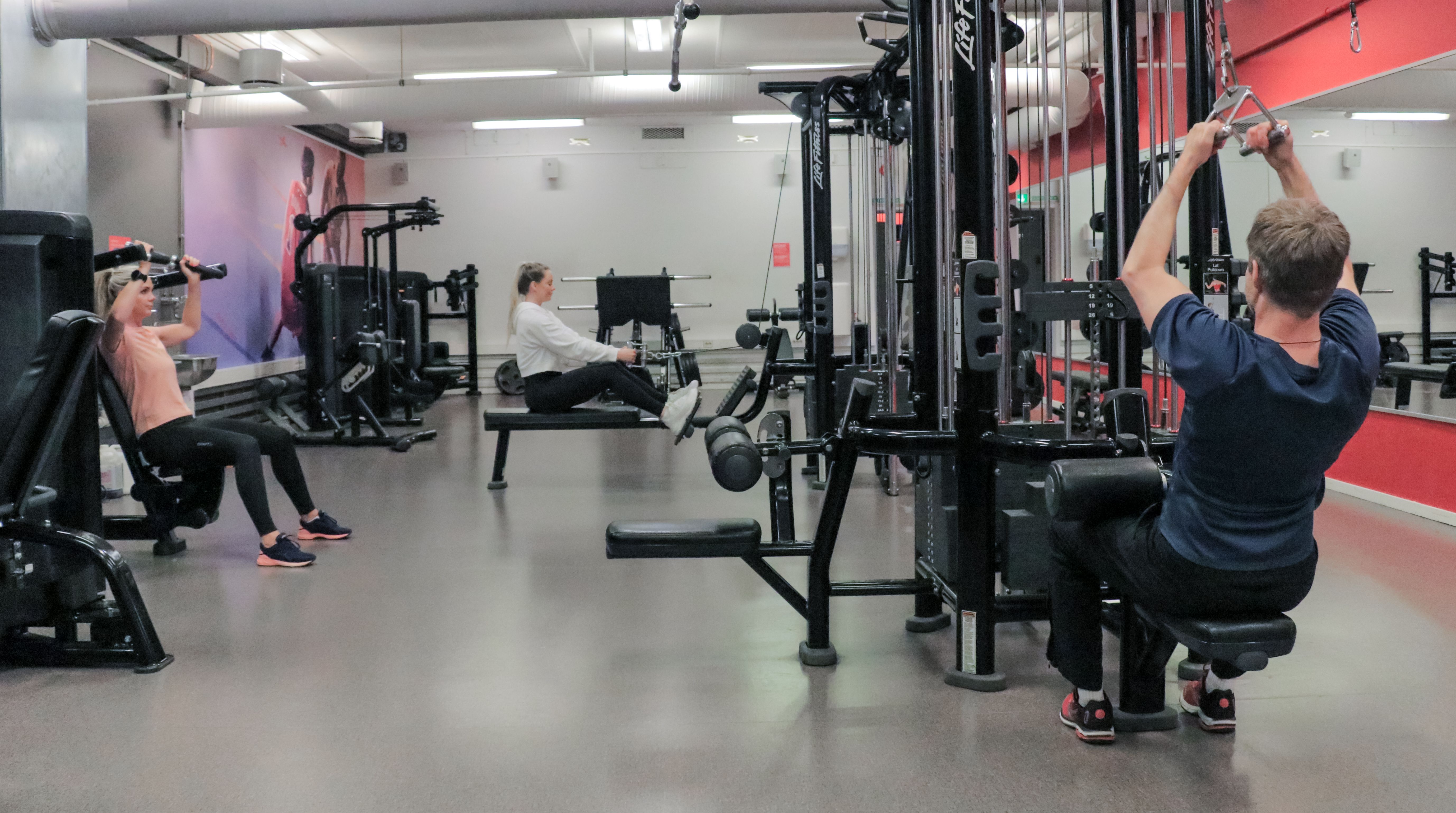 Studio på Athletica Blindern med ulike apparater og kabeltrekk, og tre personer som trener. 